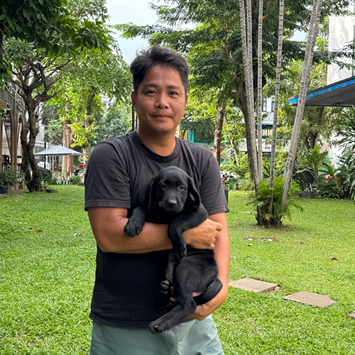 Khách hàng bế chó Labrador con màu đen chụp ngoài trời trên bãi cỏ, nền cây xanh